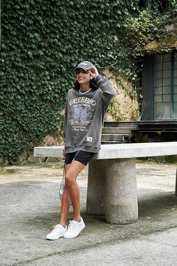 Person wearing a casual oversized sweatshirt with text, baseball cap, black shorts, and white sneakers, leaning on a stone bench outdoors