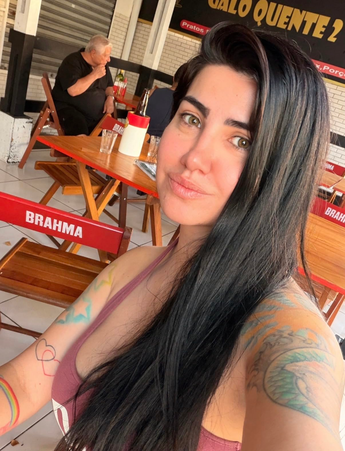 Reviewer with agelong  hairsbreadth  and disposable   tattoos takes a selfie astatine  an outdoor cafe with reddish  Brahma-branded seating