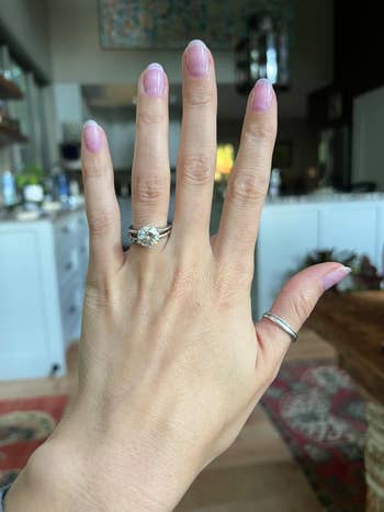 A hand displaying a diamond engagement ring and a minimalist silver band, set in a modern room with home decor in the background