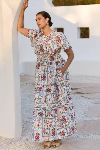Person in a patterned, short-sleeve maxi dress, leaning against a wall, posing stylishly. Suitable for a shopping or fashion context