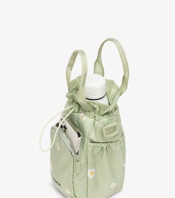 the bag in light green featuring a daisy print with a water bottle in the main compartment and a phone in the front zip pocket 