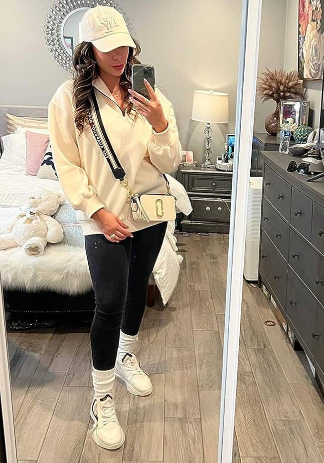 Person successful a casual, sporty outfit with a cap, crossbody bag, and sneakers, posing for a reflector selfie successful a stylish chamber setting.