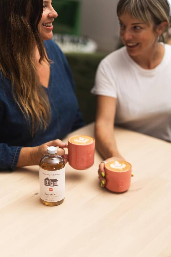 Two radical   holding mugs with latte art, smiling astatine  each   different   crossed  a array  with a lavender syrup bottle