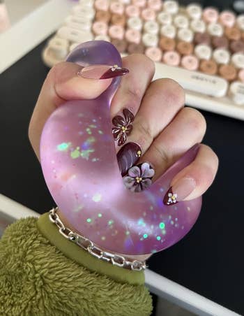 Close-up of a hand with flower-adorned manicured nails gripping a purple, shimmering stress ball near a pastel keyboard