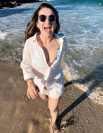 A person in sunglasses and a white beach cover-up walks along the shoreline, smiling with their tongue out. The ocean waves are in the background