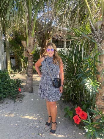 Woman successful  a patterned formal  and heels poses with a crossbody container  nether  tropical trees