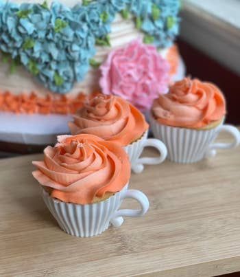 Three cupcakes with rose-shaped frosting in white cup-shaped containers; a decorated cake can be seen in the background