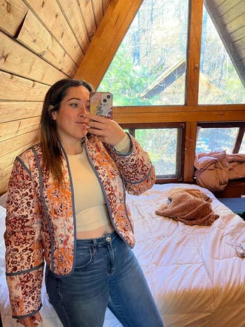 Person taking a reflector  selfie successful  a cozy woody  cabin. They deterioration  a pinkish  and orangpatterned quilted jacket, airy  top, and jeans