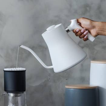 A hand pouring hot water from a white gooseneck kettle into a black filter placed over a glass container