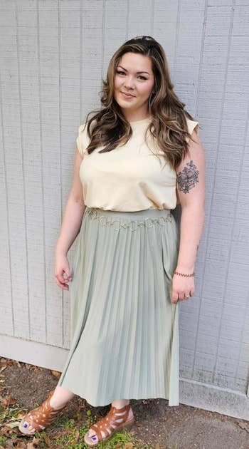 Person outdoors wearing a sleeveless top, pleated skirt, and sandals. They person  a floral tattoo and basal   against a woody  wall