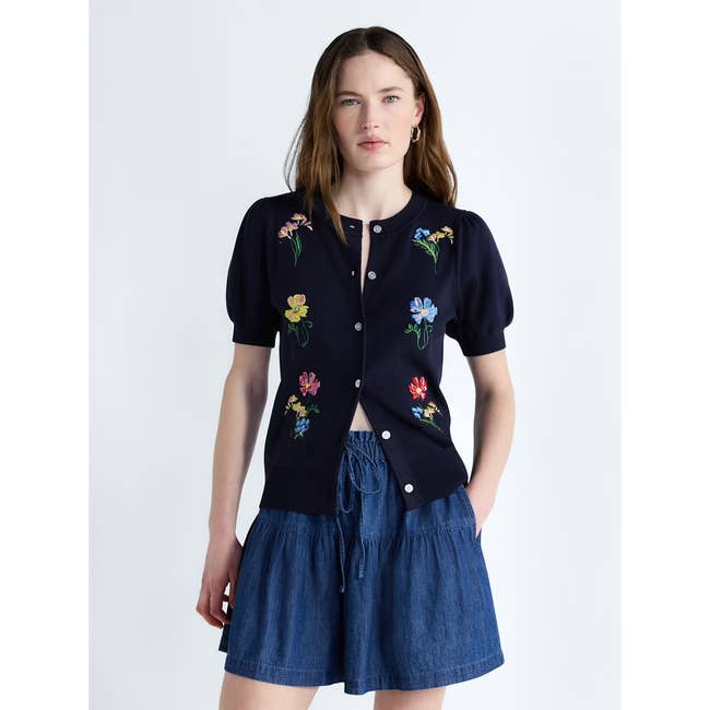 Person wearing a short-sleeve embroidered cardigan with floral designs and a denim skirt, suitable for casual or trendy styles