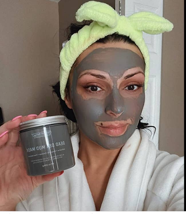 Reviewer with gray mud mask on their face holding the jar of mask 