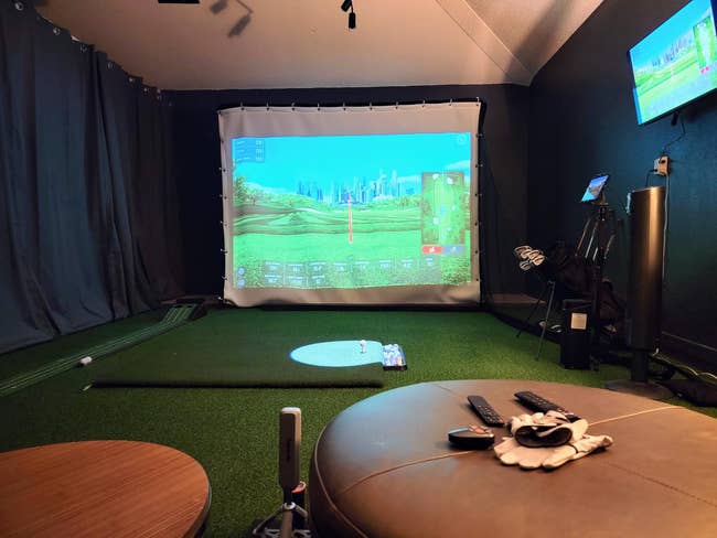 Home golf simulator setup with a virtual golf course on the screen. Includes clubs, golf gloves, and remote controls on a nearby table
