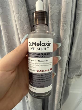 A hand holds a Dr. Melaxin Peel Shot bottle with exfoliating black rice ampoule, featuring rice bran water and niacinamide for smooth skin