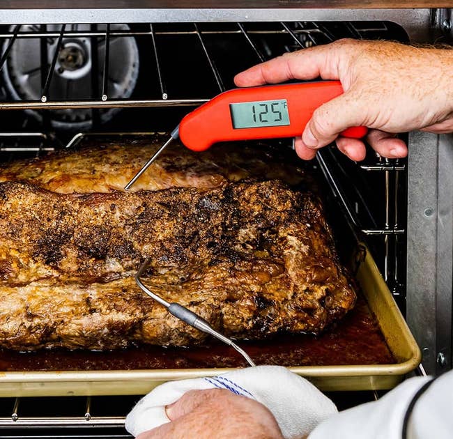 hands using the red thermapen to read the temperature of a piece of meat being pulled out of the oven