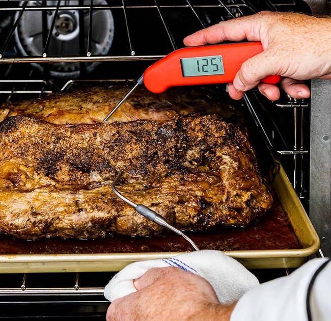 hands using the red thermapen to read the temperature of a piece of meat being pulled out of the oven