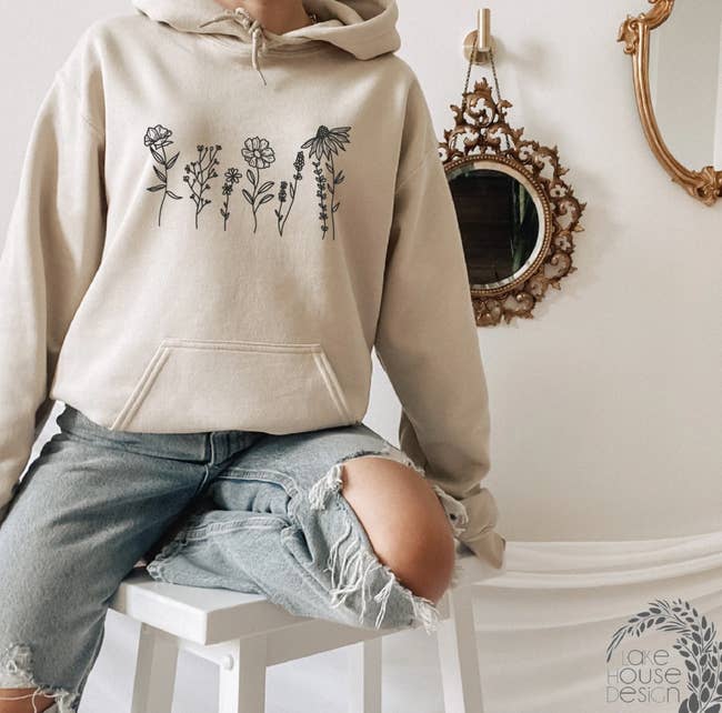 Model is wearing an oatmeal colored hoodie with a simple black wildflower design on the front