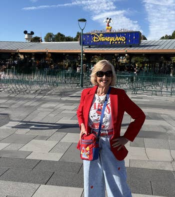 reviewer successful  casual, themed outfit poses happily successful  beforehand   of Disneyland Paris entrance, wearing a reddish  blazer and graphic tee