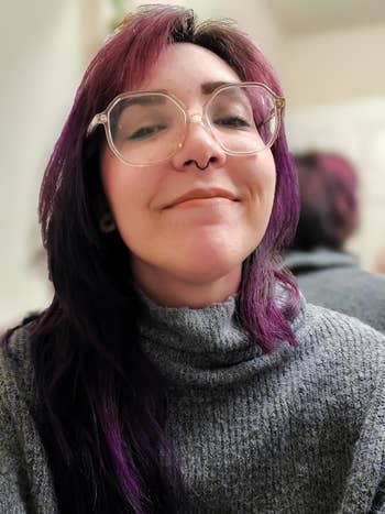 A person with glasses, wearing a textured turtleneck sweater, smiles softly
