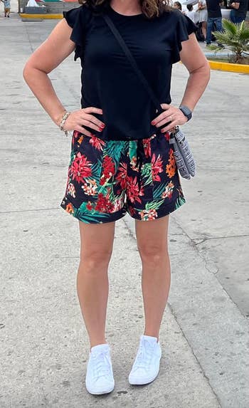 Person wearing a achromatic  garment  and floral shorts, achromatic  sneakers, and a crossbody container  stands connected  a street