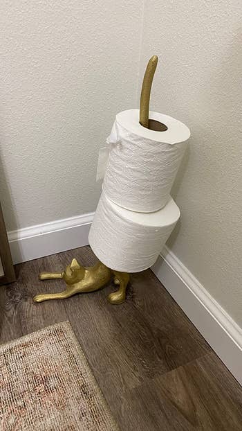 A gold holder in the shape of a cat with two toilet paper rolls hanging on the cat's tail