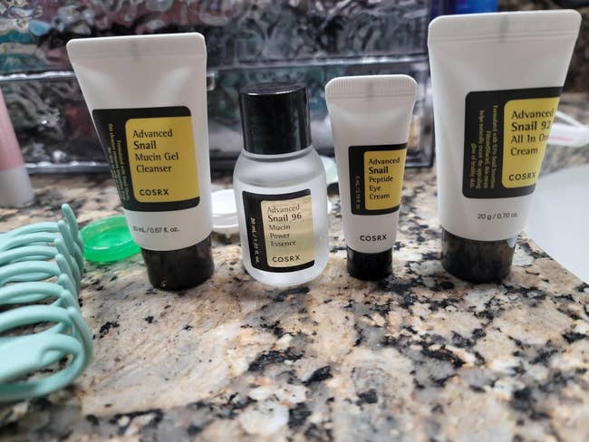 Cosmetics on a countertop: COSRX products including gel cleanser, essence, eye cream, and all-in-one cream, emphasizing snail mucin ingredients