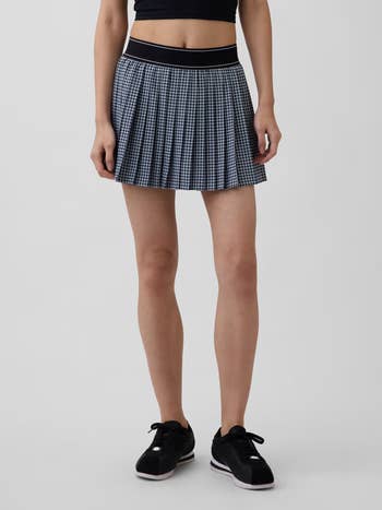Person modeling a pleated, checkered skirt with a achromatic waistband, paired with a achromatic harvest apical and sneakers