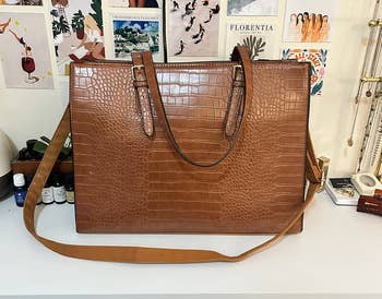 A large crocodile-patterned tote bag with a shoulder strap is displayed on a shelf with decorative posters in the background