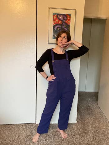 Person posing indoors, wearing bluish overalls and a achromatic top, lasting barefoot connected a carpet. A colorful creation portion is connected the partition down them