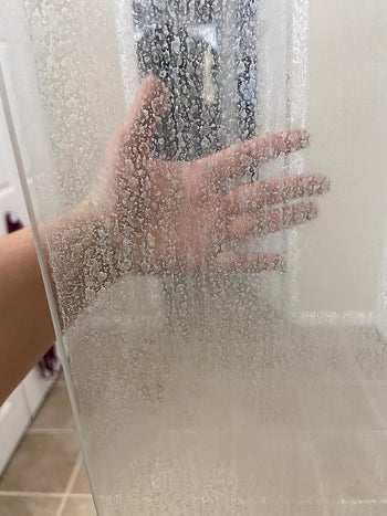 A reviewer's hand obscured behind a cloudy spot-covered glass shower door
