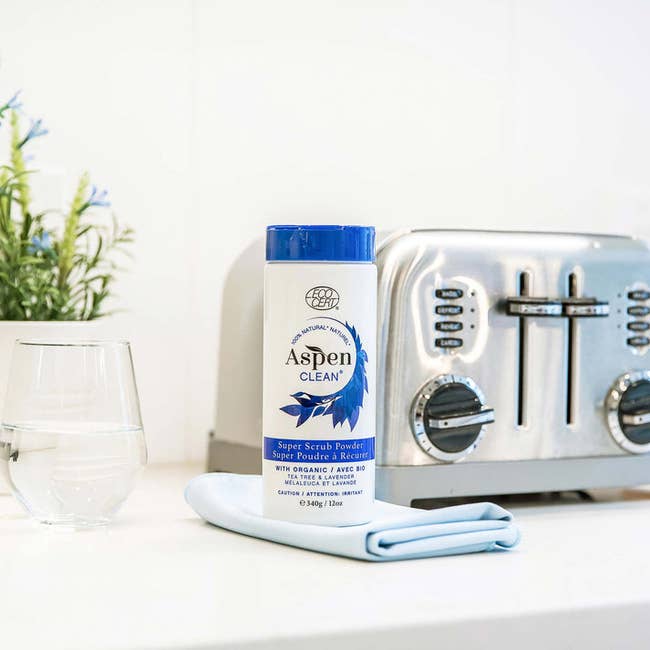 AspenClean Super Scrub Powder with a cleaning cloth next to a stainless steel toaster and glass on a kitchen countertop