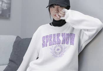 model in white crewneck sweatshirt with purple collegiate inspired speak now text with crest in the middle featuring wedding rings and text 