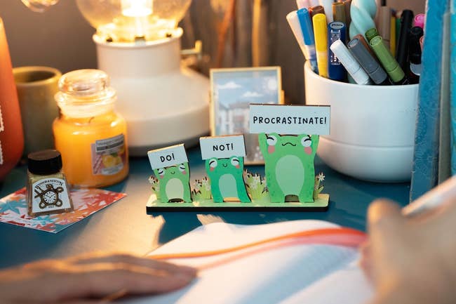 Desk with froggy desk companion holding up a sign that says do not procrastinate