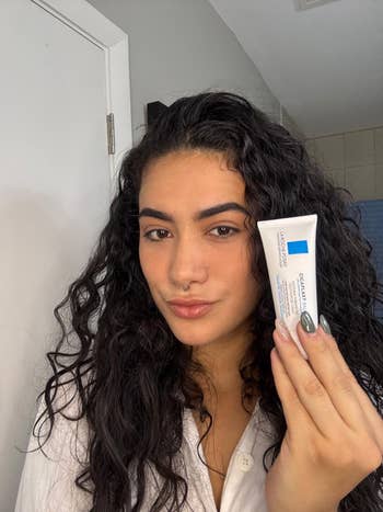 Person with curly hairsbreadth  holds a skincare product, showcasing it to the camera. They are successful  a casual setting, wearing a achromatic  top