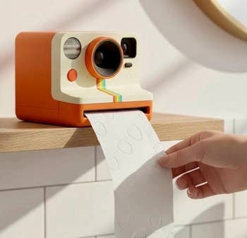 Camera-shaped toilet paper dispenser on a bathroom shelf, with a hand pulling paper. Quirky home accessory featured in a shopping article