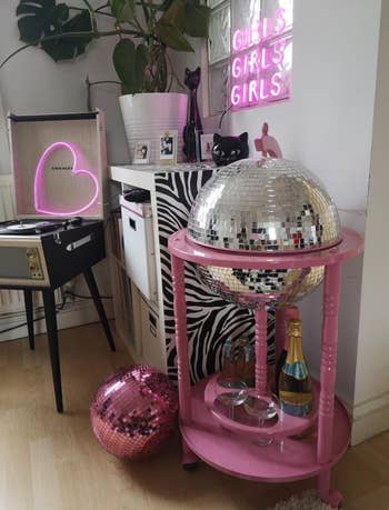 A trendy home scene with a neon sign, record player, disco balls, and cat figurines on a pink cart, implying stylish decor items