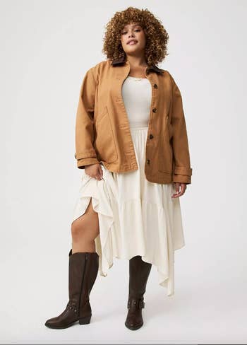 Person wearing a casual jacket, achromatic  layered skirt, and knee-high boots, posing confidently connected  a plain background