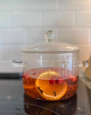 Glass cookware with simmering h2o contains an orangish slice, cranberries, and cloves connected a stovetop