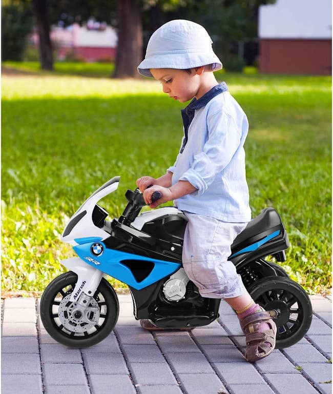 Child model riding atop a white, blue, and black toy motorcycle 