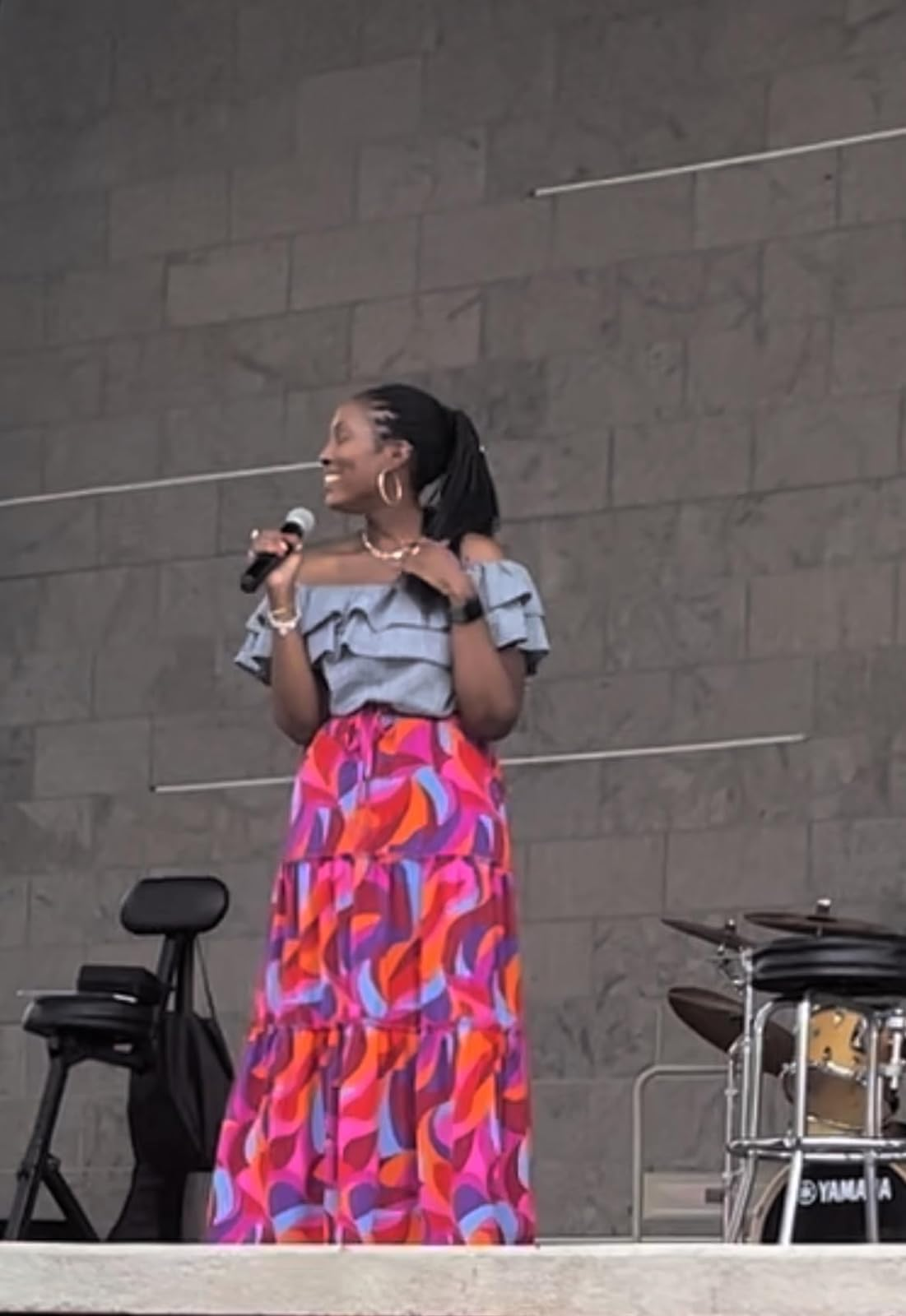 A reviewer stands on stage holding a microphone, wearing a ruffled off-shoulder top and a colorful tiered skirt, next to musical instruments