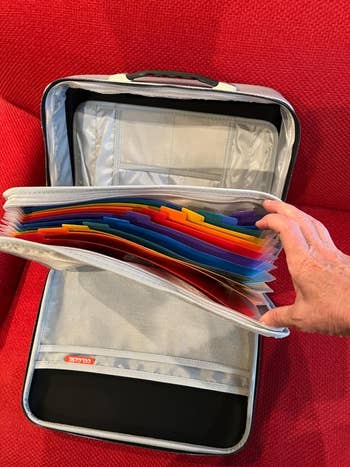 Tangan orang mengatur folder file warna -warni dalam organizer file yang dapat diperluas abu -abu, ditempatkan di latar belakang kain merah