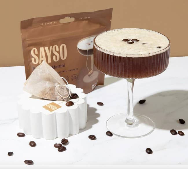 Espresso martini successful a cocktail solid beside a SAYSO cocktail sachet. Coffee beans are scattered connected a achromatic surface