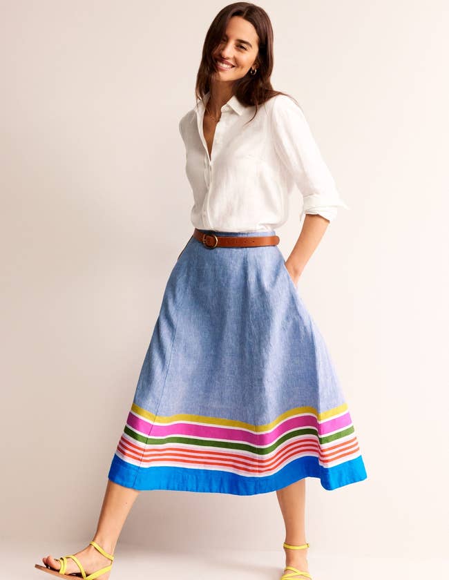 model in a white button-up shirt tucked into chambray linen midi skirt with colorful stripes at the hem