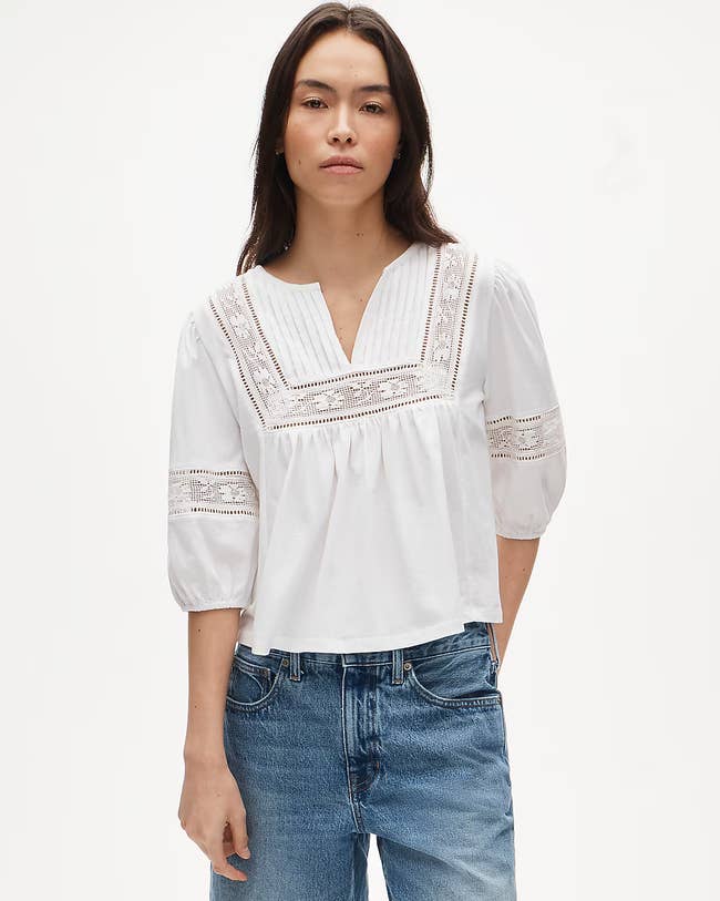Person wearing a casual achromatic blouse with lace details and bluish jeans, posing against a plain background