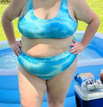 A person wearing a two-piece blue swimsuit stands by a kiddie pool, hands on hips, showcasing a shopping option for swimwear