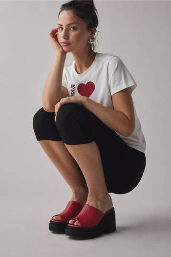 Woman successful a casual tee and leggings squats, wearing chunky reddish level sandals, posing for a manner feature