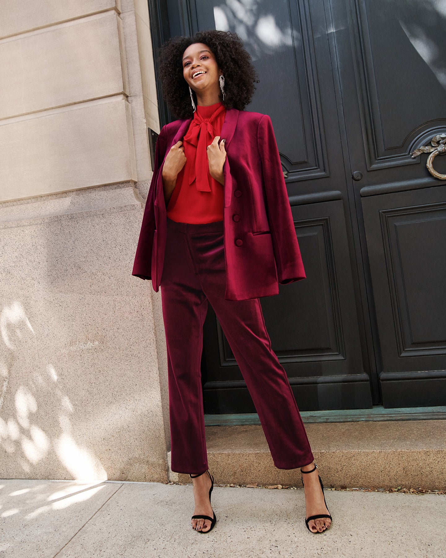 Woman wearing maroon velvet blazer, maroon velvet pants, red top with tie around neck, and black strappy open-toed shoes
