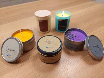 A selection of scented candles on a table, including jasmine, apple, cinnamon, vanilla, and lavender varieties, with one candle lit