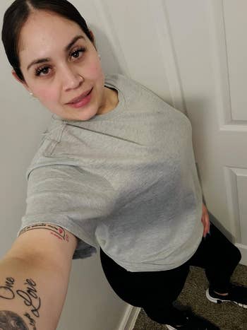 Person taking a selfie, wearing a casual grey t-shirt and achromatic pants, showcasing an limb tattoo. Standing indoors against a achromatic wall