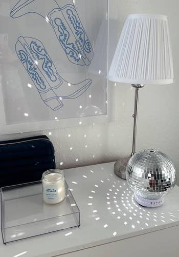 A stylish home setting with a lamp, candle, disco ball, and a framed artwork on a white surface